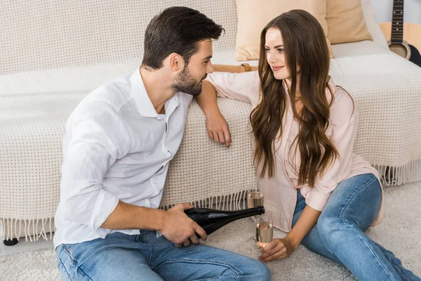 Homme verser du champagne dans le verre tout en étant assis sur le sol avec sa petite amie à la maison — Photo de stock