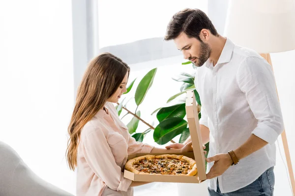 Vista lateral de pareja joven mirando pizza delivired en caja de papel en casa - foto de stock