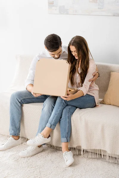 Coppia sorridente con pizza in scatola di carta seduta sul divano di casa — Foto stock