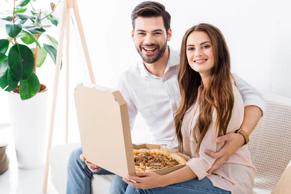 Portrait de couple souriant avec pizza dans une boîte en papier assise sur un canapé à la maison — Photo de stock