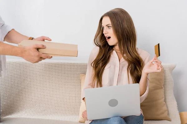 Vista parcial del hombre con pizza en caja de papel y mujer sorprendida con tarjeta de crédito y portátil en casa, concepto de compras en línea - foto de stock