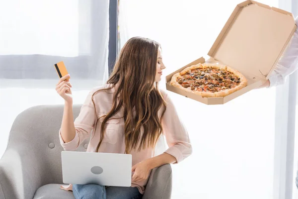 Vista parcial de hombre con pizza en caja de papel y mujer joven con tarjeta de crédito y portátil en casa, concepto de compras en línea - foto de stock