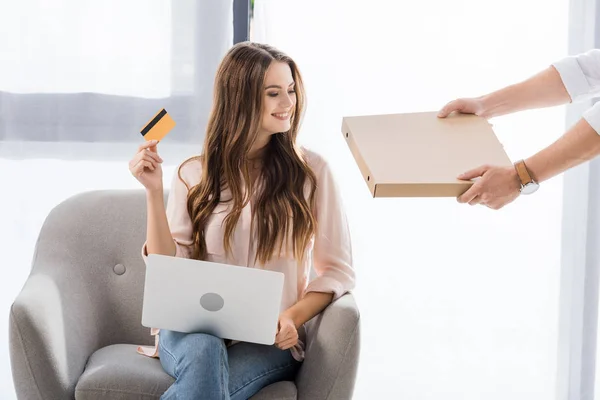 Vista parziale dell'uomo con pizza in scatola di carta e donna sorridente con carta di credito e laptop a casa, concetto di shopping online — Foto stock