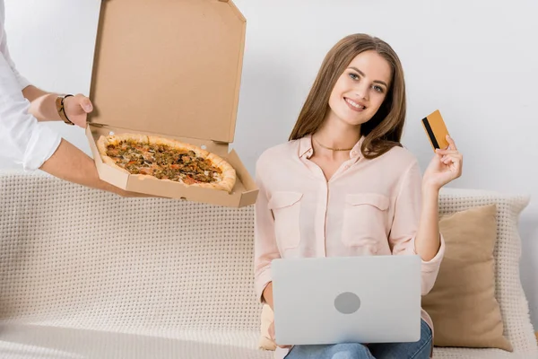 Visão parcial do homem com pizza em caixa de papel e mulher sorridente com cartão de crédito e laptop em casa, compras conceito on-line — Fotografia de Stock