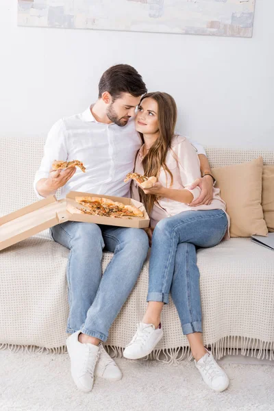 Pareja joven con pizza en caja de papel descansando en el sofá en casa - foto de stock