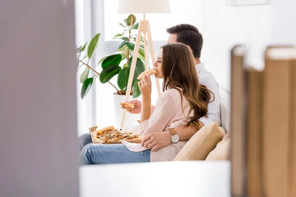 Vue latérale du jeune couple mangeant de la pizza sur le canapé à la maison — Photo de stock