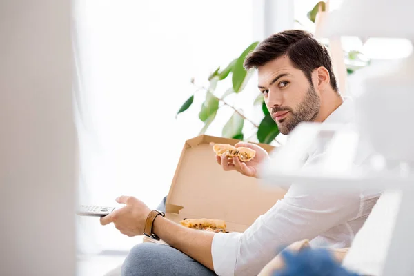 Seitenansicht eines jungen Mannes mit Fernbedienung in der Hand, der allein zu Hause Pizza isst — Stockfoto