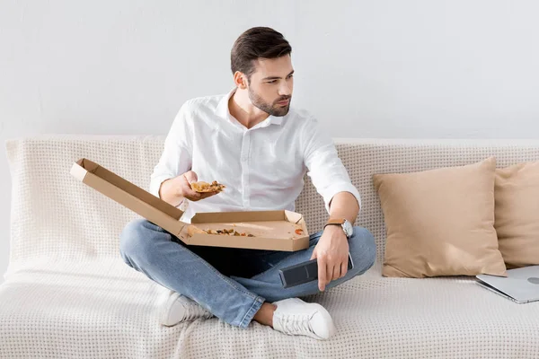 Молодой человек с пультом дистанционного управления в руке едят пиццу в одиночестве дома — стоковое фото