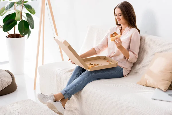 Young smiling woman eating pizza while resting on sofa at home — Stock Photo