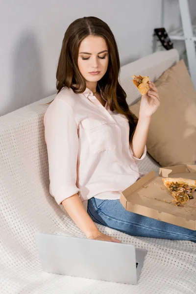 Young woman with pizza using laptop while resting on sofa at home — Stock Photo