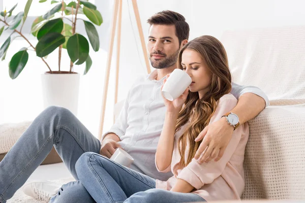Young loving couple with cups of coffee on sofa at home — Stock Photo