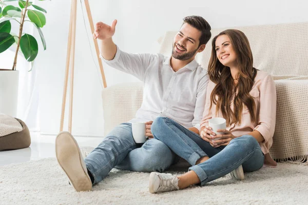 Happy loving couple with cups of coffee sitting on floor and watching tv at home — Stock Photo
