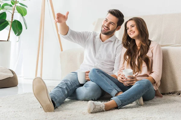 Feliz casal amoroso com xícaras de café sentado no chão e assistir tv em casa — Fotografia de Stock