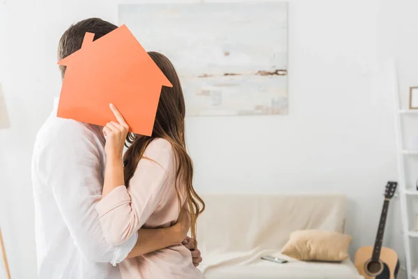 Obscured view of couple kissing behind paper house model in hand at new home — Stock Photo