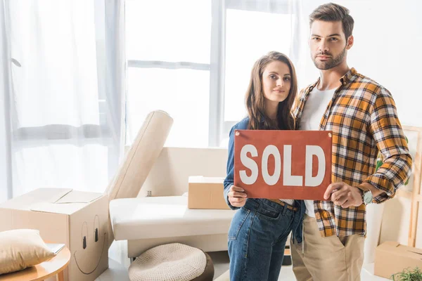 Portrait of young couple holding sold red card at home with cardboard boxes — Stock Photo