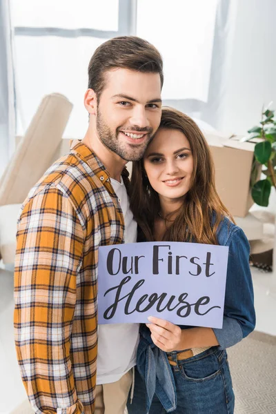 Portrait of smiling couple with our first house card standing in room with cardboard boxes — Stock Photo