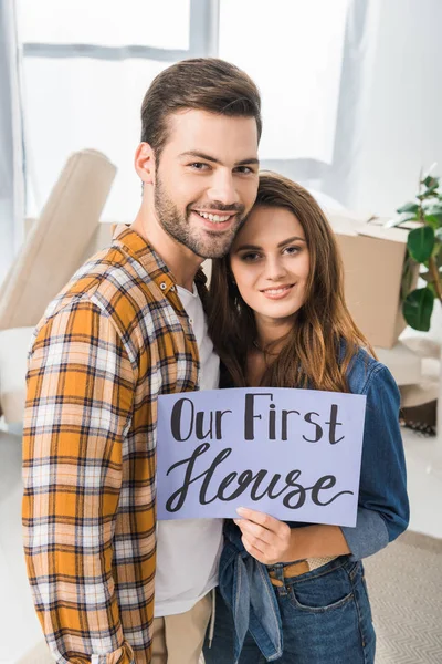 Retrato de pareja sonriente con nuestra primera tarjeta de la casa de pie en la habitación con cajas de cartón - foto de stock