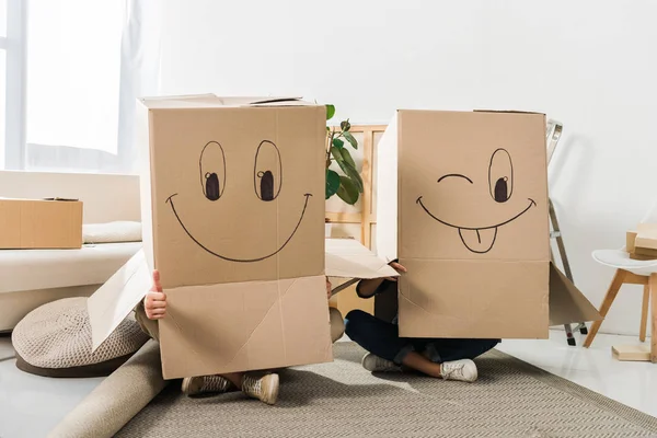 Vista oscurecida de pareja con cajas de cartón sobre cabezas sentadas en el piso en casa nueva, concepto de casa móvil - foto de stock