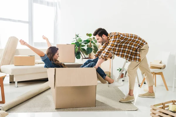 Vista lateral do casal se divertindo com caixa de papelão na nova casa, movendo-se para casa conceito — Fotografia de Stock