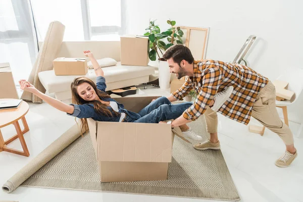Vista lateral do casal se divertindo com caixa de papelão na nova casa, movendo-se para casa conceito — Fotografia de Stock