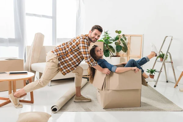 Вид сбоку улыбающейся пары, веселящейся с картонной коробкой в новом доме, концепция переезда домой — стоковое фото
