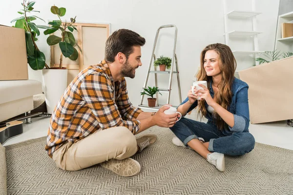 Lächelndes Paar mit Kaffeebecher auf dem Boden im neuen Haus sitzend, Umzugskonzept — Stockfoto