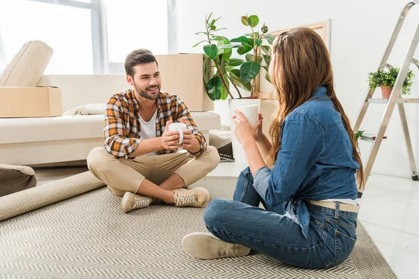 Jovem casal com xícaras de café sentado no chão em casa nova, movendo-se conceito de casa — Fotografia de Stock