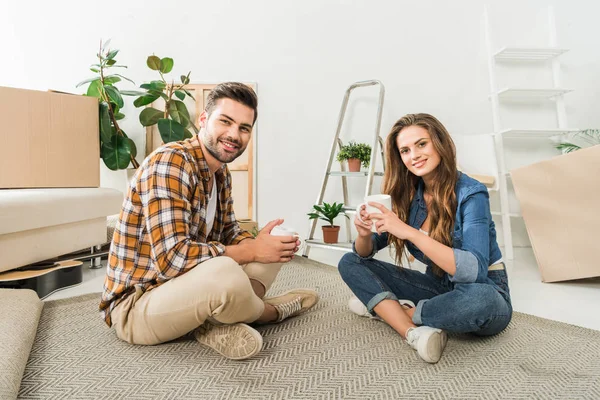 Smiling couple with cups of coffee sitting on floor at new house, moving home concept — Stock Photo