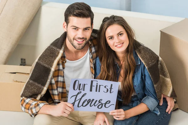 Portrait of smiling couple in blanket with our first house card sitting on sofa, moving home concept — Stock Photo