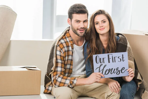 Portrait of young couple in blanket with our first house card sitting on sofa, moving home concept — Stock Photo