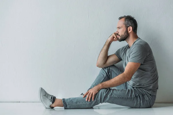 Vista lateral del frustrado hombre barbudo de mediana edad sentado en el suelo y mirando hacia otro lado - foto de stock