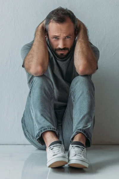 Hombre deprimido asustado sentado en el suelo con las manos detrás de la cabeza y mirando a la cámara - foto de stock