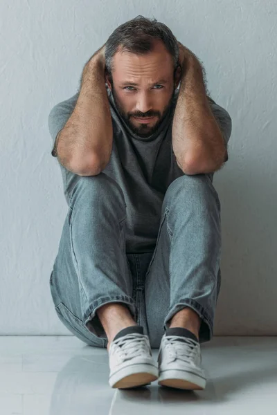 Homme déprimé effrayé assis sur le sol avec les mains derrière la tête et regardant la caméra — Photo de stock