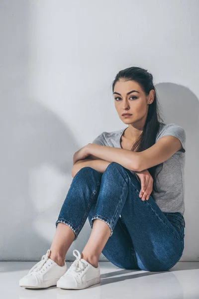 Volle Länge Ansicht der traurigen jungen brünetten Frau sitzt auf dem Boden und schaut in die Kamera — Stockfoto