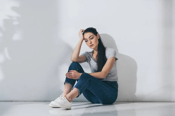 Unhappy young brunette woman with hand on forehead sitting on floor near grey wall — Stock Photo