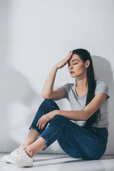 Jeune femme malheureuse avec les yeux fermés et la main sur le front assis sur le sol près du mur gris — Photo de stock