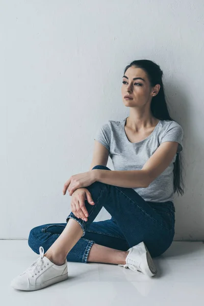 Depressive junge brünette Frau sitzt in der Nähe einer grauen Wand und schaut weg — Stockfoto