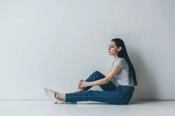 Side view of frustrated young brunette woman sitting near grey wall and looking away — Stock Photo