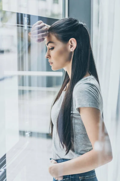Side view of upset young woman leaning at window and looking down — Stock Photo