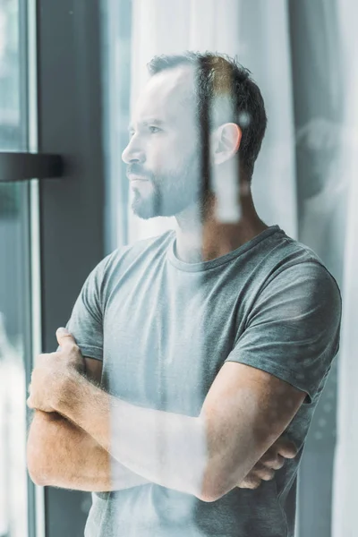 Селективное внимание бородатого мужчины средних лет, стоящего со скрещенными руками и смотрящего в окно — стоковое фото