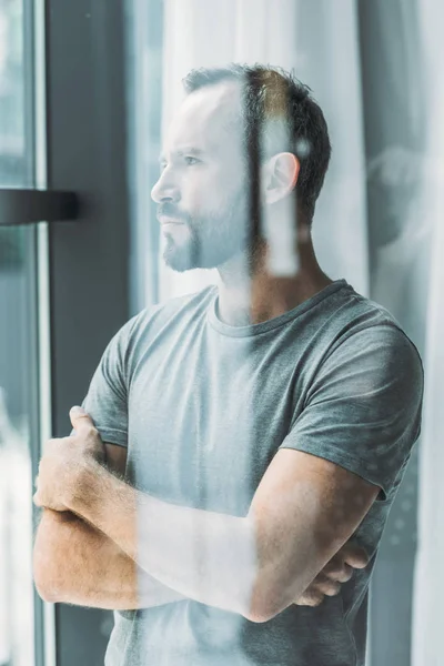 Enfoque selectivo del hombre barbudo de mediana edad de pie con los brazos cruzados y mirando a la ventana - foto de stock