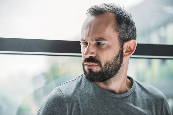 Portrait d'un homme barbu malheureux debout près de la fenêtre et regardant ailleurs — Photo de stock