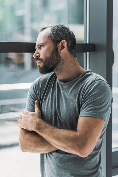 Sad bearded middle aged man standing with crossed arms and looking at window — Stock Photo