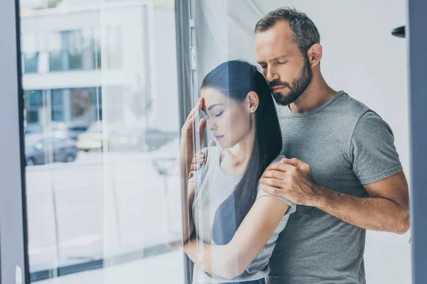 Bearded man supporting sad stressed woman with closed eyes standing near window — Stock Photo
