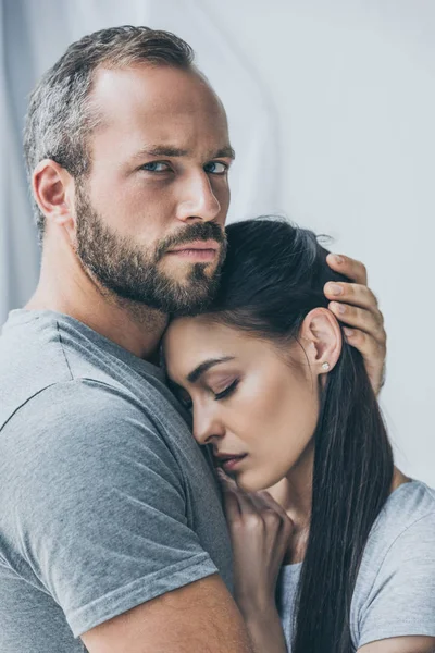Bärtiger Mann umarmt aufgebrachte Freundin und blickt in Kamera — Stockfoto
