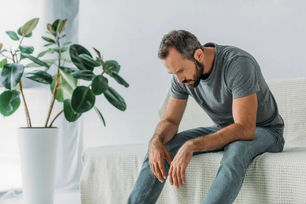 Депрессивный бородатый мужчина среднего возраста сидит на диване и смотрит вниз — стоковое фото