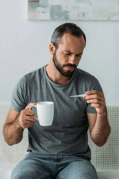 Kranker bärtiger Mann mittleren Alters mit Tasse und Blick auf Thermometer — Stockfoto