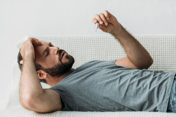 Kranker bärtiger Mann liegt auf Couch und hält Thermometer — Stockfoto