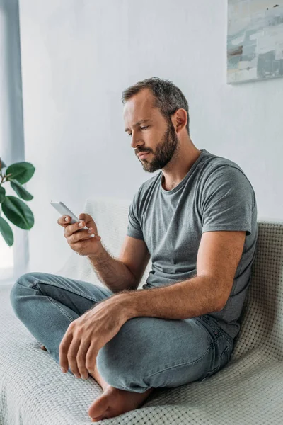 Infeliz hombre barbudo de mediana edad sentado en el sofá y el uso de teléfono inteligente - foto de stock