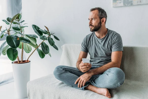 Hombre barbudo molesto sosteniendo el teléfono inteligente y mirando hacia otro lado mientras está sentado en el sofá en casa - foto de stock